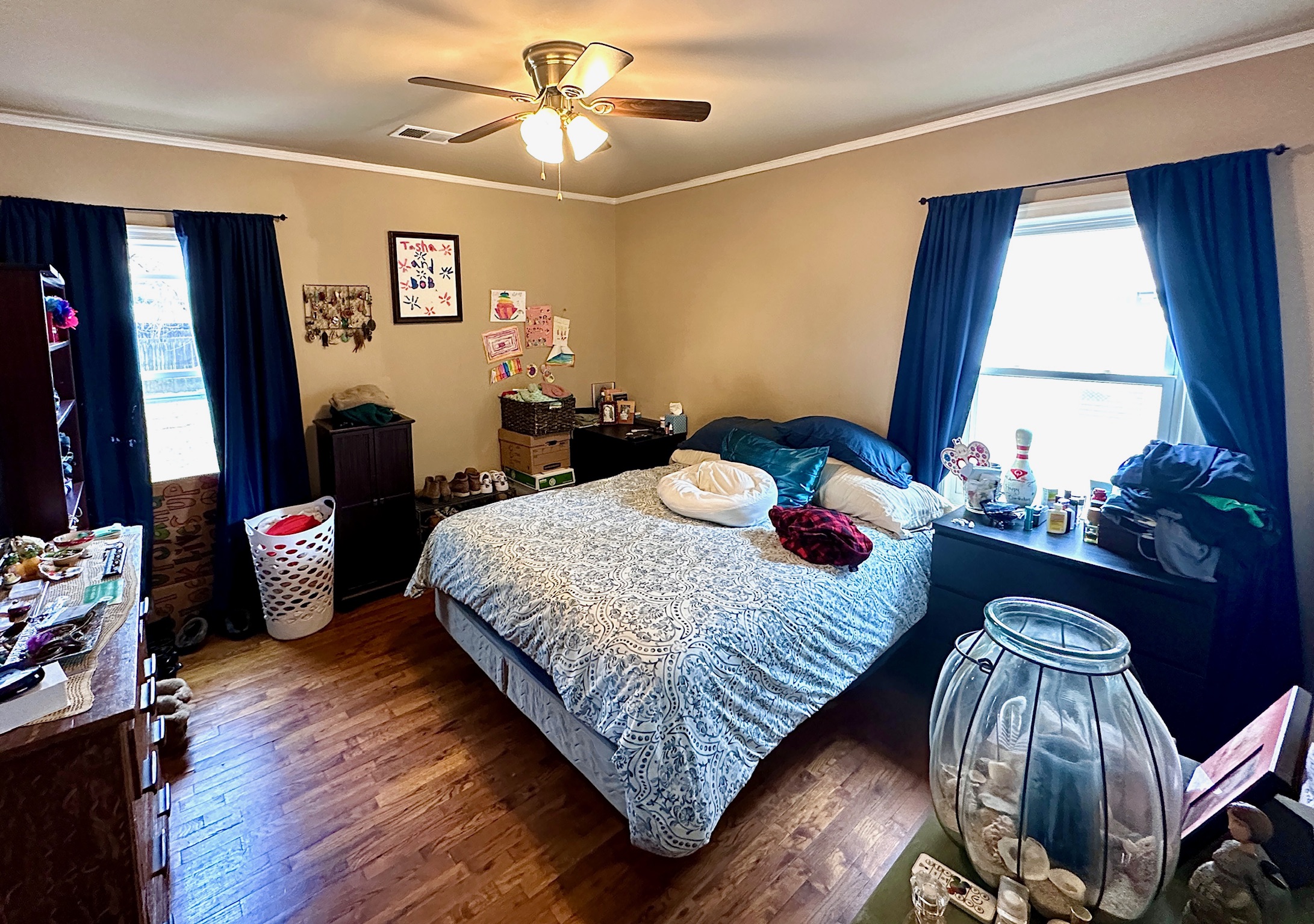 A cozy bedroom featuring a bed with pillows, dressers, and a window with curtains.